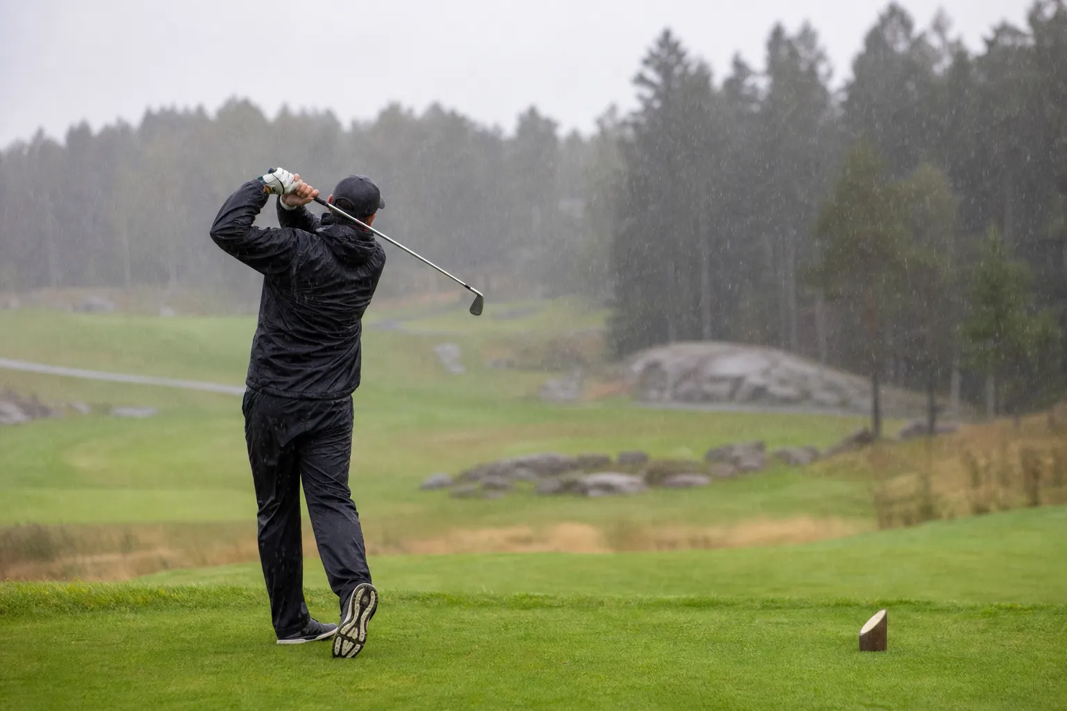 En golfare i regnjacka och regnbyxor mitt i en sving på en regnig golfbana